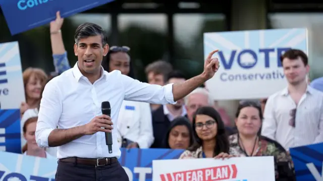 Rishi Sunak campaigns in a white dress shirt in front of a crowd of people holding signs for the Conservative Party.