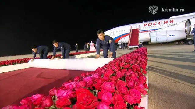 Three men roll out a red carpet in between boxes containing hundreds of red roses