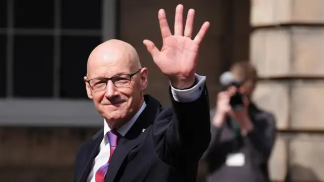 John Swinney raises his hand in a wave to the camera