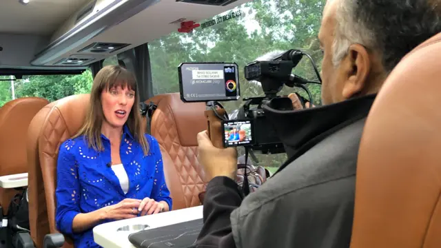 An image showing Jenny Hill sitting at a table on board the Lib Dems' campaign bus and talking, while she is being filmed by a colleague.