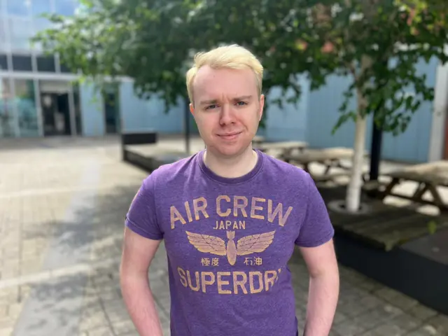 A young man with bleached blonde hair stands in a sunlit courtyard, hands on his hips. He's wearing a purple t-shirt with "Air Crew, Japan, Superdry" printed on it in yellow-gold lettering.