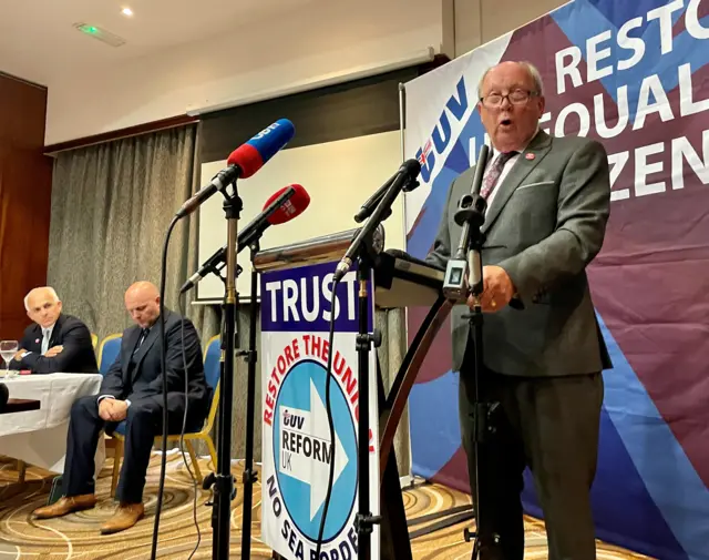 An image showing TUV leader wearing a suit and standing behind a lectern while giving a speech