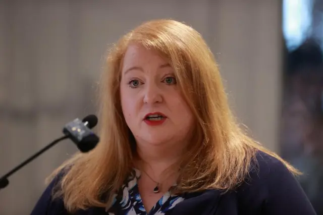 Naomi Long,  wearing a blue cardigan,  speaking into a microphone