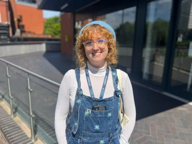 A young woman stands in front of a glass-fronted building, smiling. She's wearing a white, long-sleeved polo neck under denim dungarees with small blue flowers embroidered on them. She has glasses, curly red hair and wears a light blue beret.