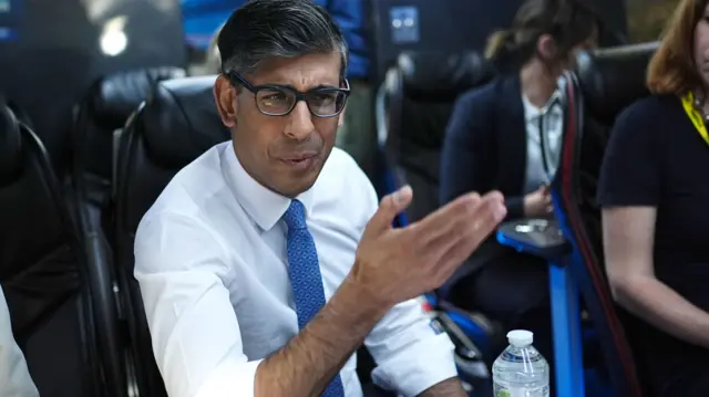 Prime Minister Rishi Sunak sits at a table on the Conservatives' battle bus and speaks to reporters