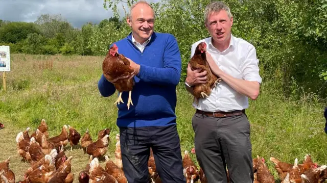 Sir Ed Davey holds a chicken while standing next to a man in white dress shirt.