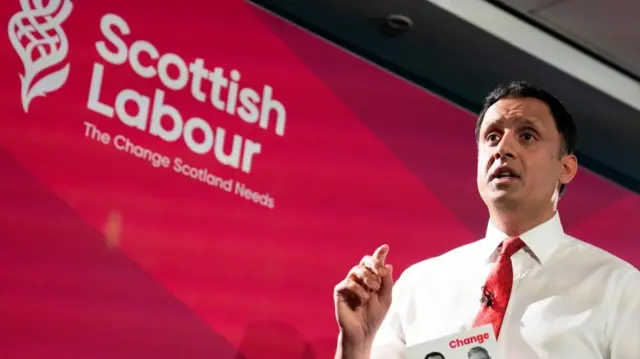 Scottish Labour leader Anas Sarwar standing in front of a red background with the words Scottish Labour, mid-speech with one hand gesturing in the air and the other holding his party's manifesto brochure.