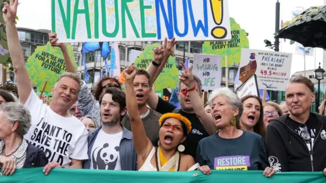 A group of people hold up a large banner in front of them at a march, with several holding up one arm in the air and others in the background holding up protest signs