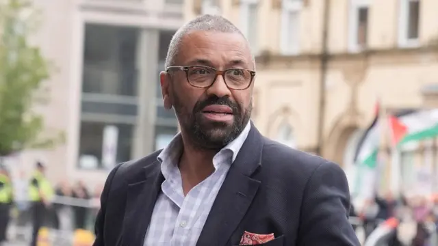 UK Home Secretary James Cleverly in a dark grey suit pictured outside during the day with a blurred background of people and buildings behind him