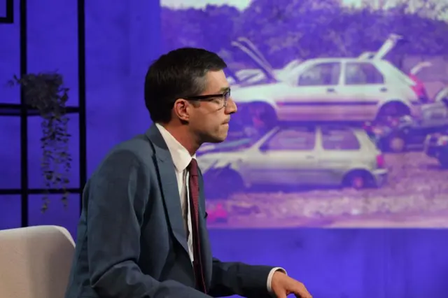 Side profile of Green Party co-leader Adrian Ramsay sat in front of a picture of written off cars