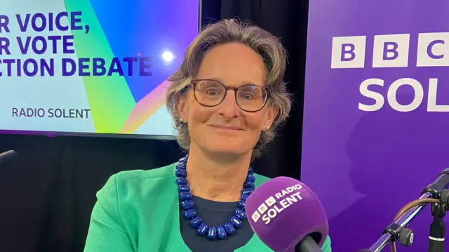 Flick Drummond in a green jacket with blue necklace, sitting in front of a microphone