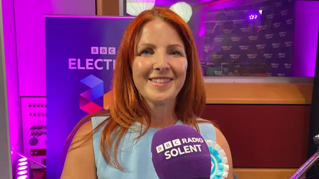 Sarah Morris in a light blue dress, sitting in front of a microphone