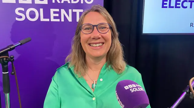 Netty Shepherd in a green blouse, sitting in front of a microphone