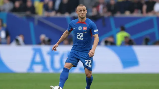 Slovakia midfielder Lobotka competing against Romania during Euro 2024.  He is standing with the ball on his right foot.  He's wearing a blue jersey with number 22 in white