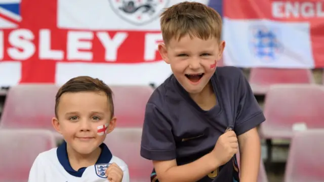 Two young England fans proudly showing off their England badges on their shirts.  They are in a stadium in Germany preparing to watch their team play at Euro 2024.  Both boys have a look of happiness and excitement