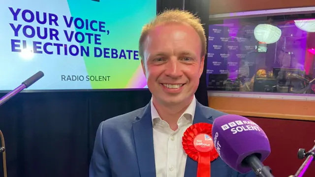 Darren paffey in a navy blue suit with red rosette, sitting in front of a microphone