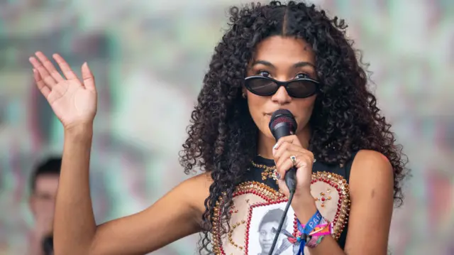 Singer Olivia Dean peers through her sunglasses, with a hand in the air as she performs on the Pyramid Stage.