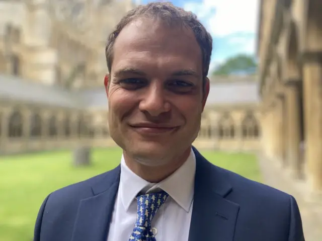 Hamish Falconer in a suit and tie smiling in the grounds of Lincoln Cathedral