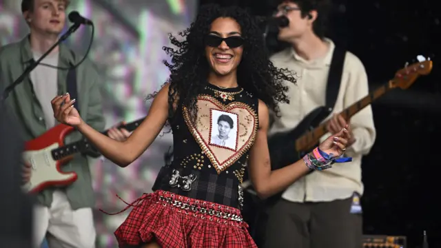 Singer Olivia Dean smiles as she performs on the Pyramid Stage, while wearing sunglasses and a top that features a photo of her grandmother inside a heart.