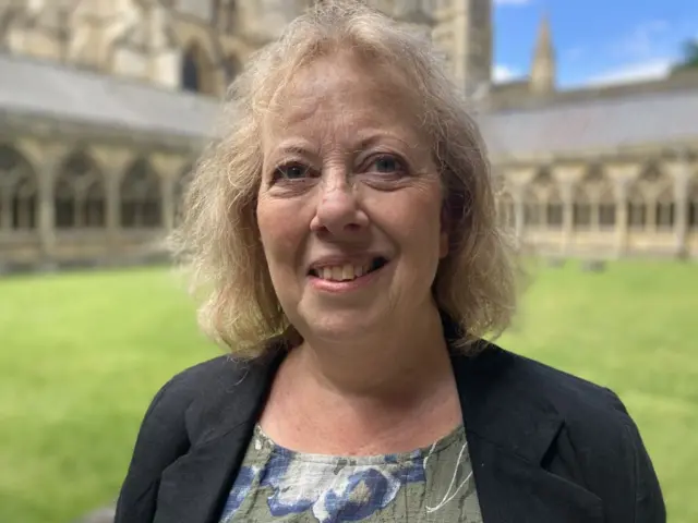 Sally Horscroft smiling with a black blazer and flower top on in the grounds of Lincoln Cathedral