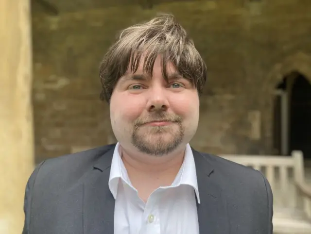 Jamie-Lee McMillan in a suit with his top button undone in the grounds of Lincoln Cathedral