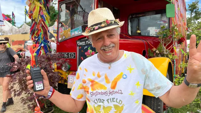 A man standing with his arms aloft in front of a big red bus. He's holding a walkie-talkie two way radio in his right hand and is wearing a colourful straw hat.