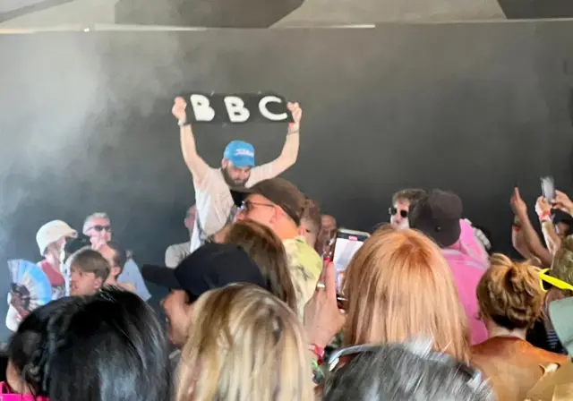 A man holds up a BBC sign while dancing in the crowd at Glastonbury.