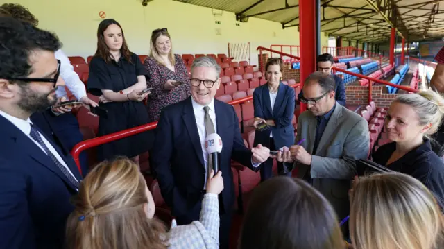 Starmer stands surrounded by the media. A woman holds a LBC microphone is held in front of him.