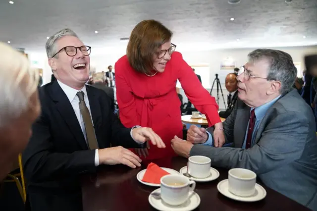 Labour Party leader Sir Keir Starmer laughing while seated at a small table with several tea cups, with Labour candidate Alex Baker standing next time him talking to a man also seated at the table