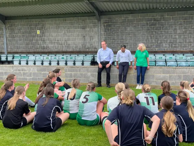 Wide shot showing the backs of about two dozen young women in football kits seated on a football pitch, looking at Prime Minister Rishi Sunak who is seated on a raised wall with stadium seats behind him