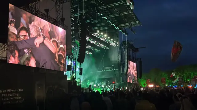 A screen next to a stage shows a National band member hugs the crowd