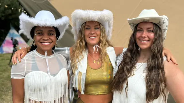 three female dancers wearing cowboy hats and tassle covered tops while standing behind the scenes of the Pyramid Stage at Glastonbury Festival.