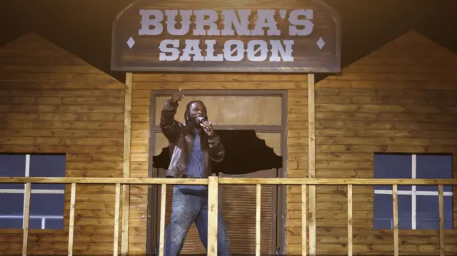 Burna Boy stands in front of a stage backdrop that looks like a wooden saloon with a sign above saying Burna's Saloon