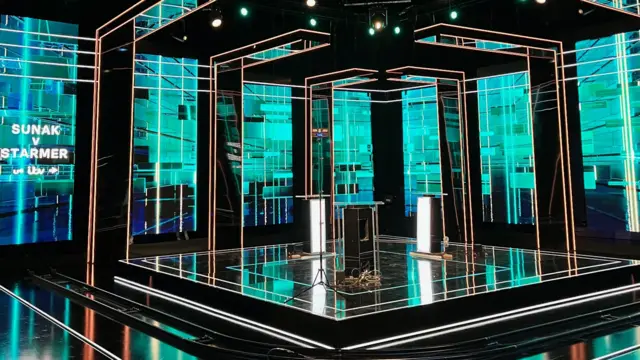 Wide shot of inside the TV studio where the debate will happen, with shiny black floors, blue-green lighting, the speakers' podiums and a neon sign saying "Sunak v Starmer"