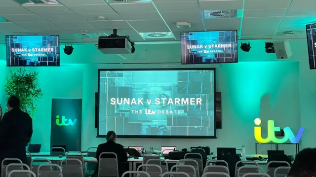 Rows of chairs facing a sign projected on to the wall saying Sunak v Starmer lit in turquoise light