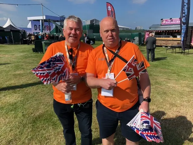 Two men in orange t-shirts holding numerous union flags on Southsea Common