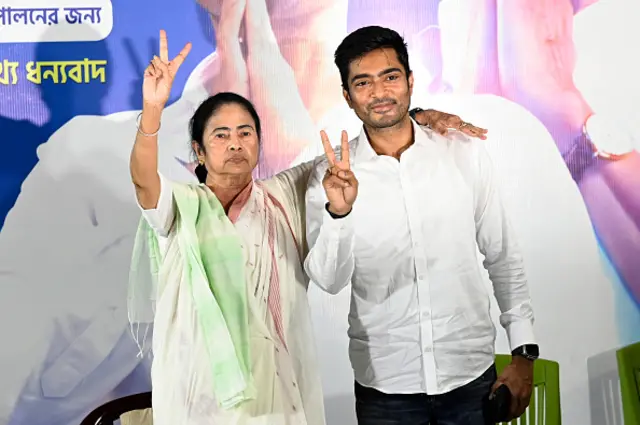 Mamata Banerjee, seen wearing a white sari, making a victory sign with her right hand. Her left hand is around the shoulders of nephew Abhishek Banerjee who has become an MP from West Bengal