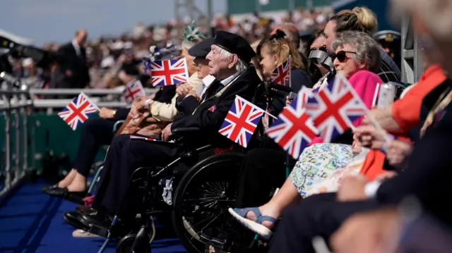 A veteran in a wheelchair watches as people around him wave Union Flags