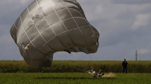 Military person lands on a green field, kicking up dirt as they land, with an open parachute billowing above them.