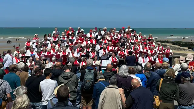 Choir dressed in white with red scarves sings while crowd looks on