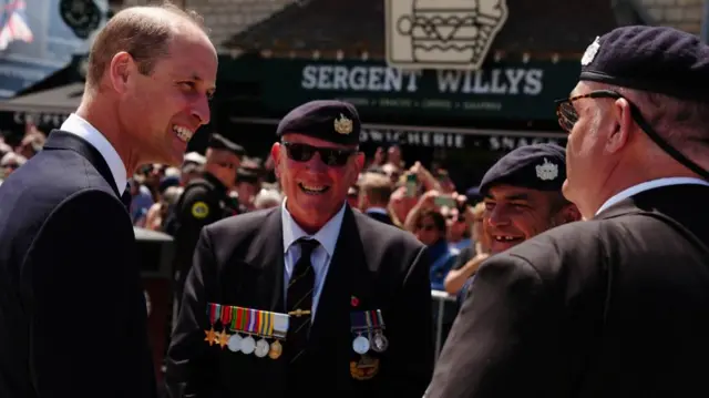 William smiles as he speaks to three veterans. In the background, a crowd has gathered by a pub called Sergent Willys