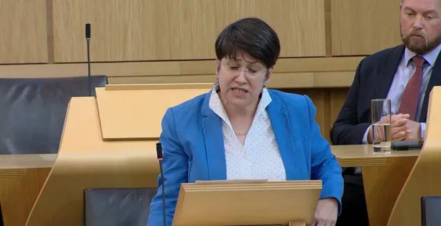 Tess White MSP stands at a lectern wearing a bright blue suit jacket and white blouse with black polka dots, she is looking down towards the lectern at papers as she speaks