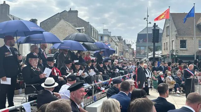 D-Day veterans sit under black umbrellas on a raised platform next to a crowd