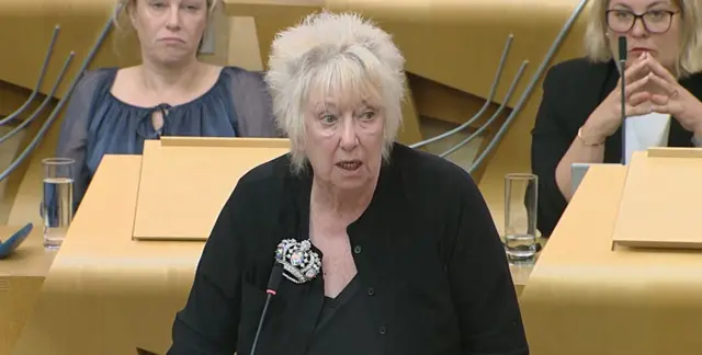 Christine Grahame MSP standing at a lectern, speaking, wearing a black cardigan, looking to her left towards Kate Forbes