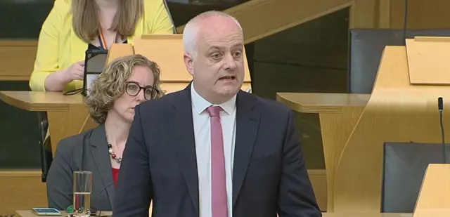 Mark Ruskell MSP wearing a navy suit and salmon pink tie, standing at a lectern speaking, looking to his left towards Kate Forbes. Green party co-leader Lorna Slater is seated behind his right shoulder.
