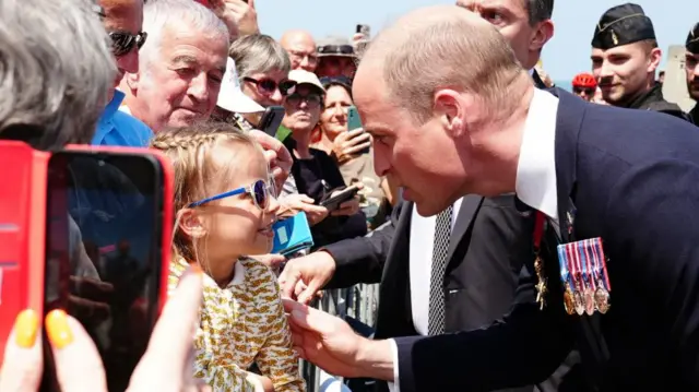 Prince William, with medals on his lapel, speaks to a little girl in sunglasses among a crowd