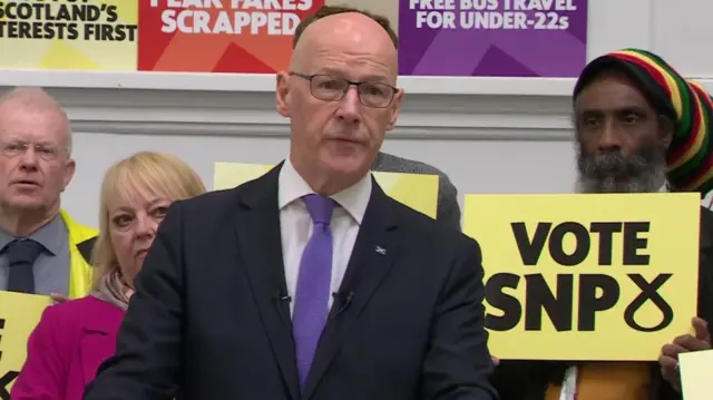 Swinney speaks. Behind him, supporters hold Vote SNP placards