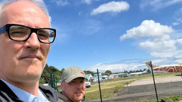 A man wearing glasses and another behind him wearing a hat looks into the camera, an airfield with planes is behind them in the distance