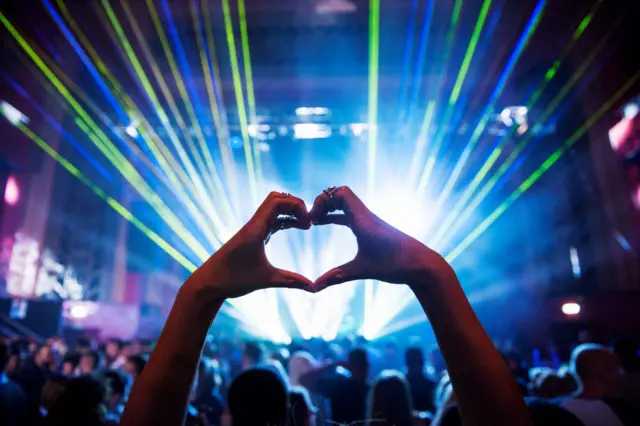 Two hands forming the shape of a heart in a dark concert venue with strobe lights shining and a large crowd facing the stage