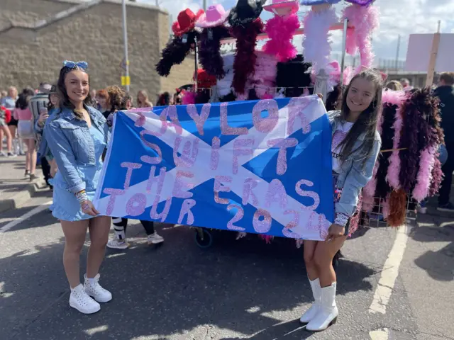 Two women holding a Saltire flag with text handwritten on in pink glitter which reads 'Taylor Swift the Eras tour 2024' - behind the girls are concert goers and a stand selling feather boas and cowboy hats